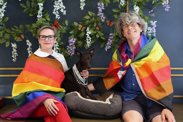 A man and a women sitting on a counter in the stephen einhorn showrrom. They have a black whippet between them who is wearing a pearl necklace. They are both draped in the colourful Pride flag. They look very cheerful.